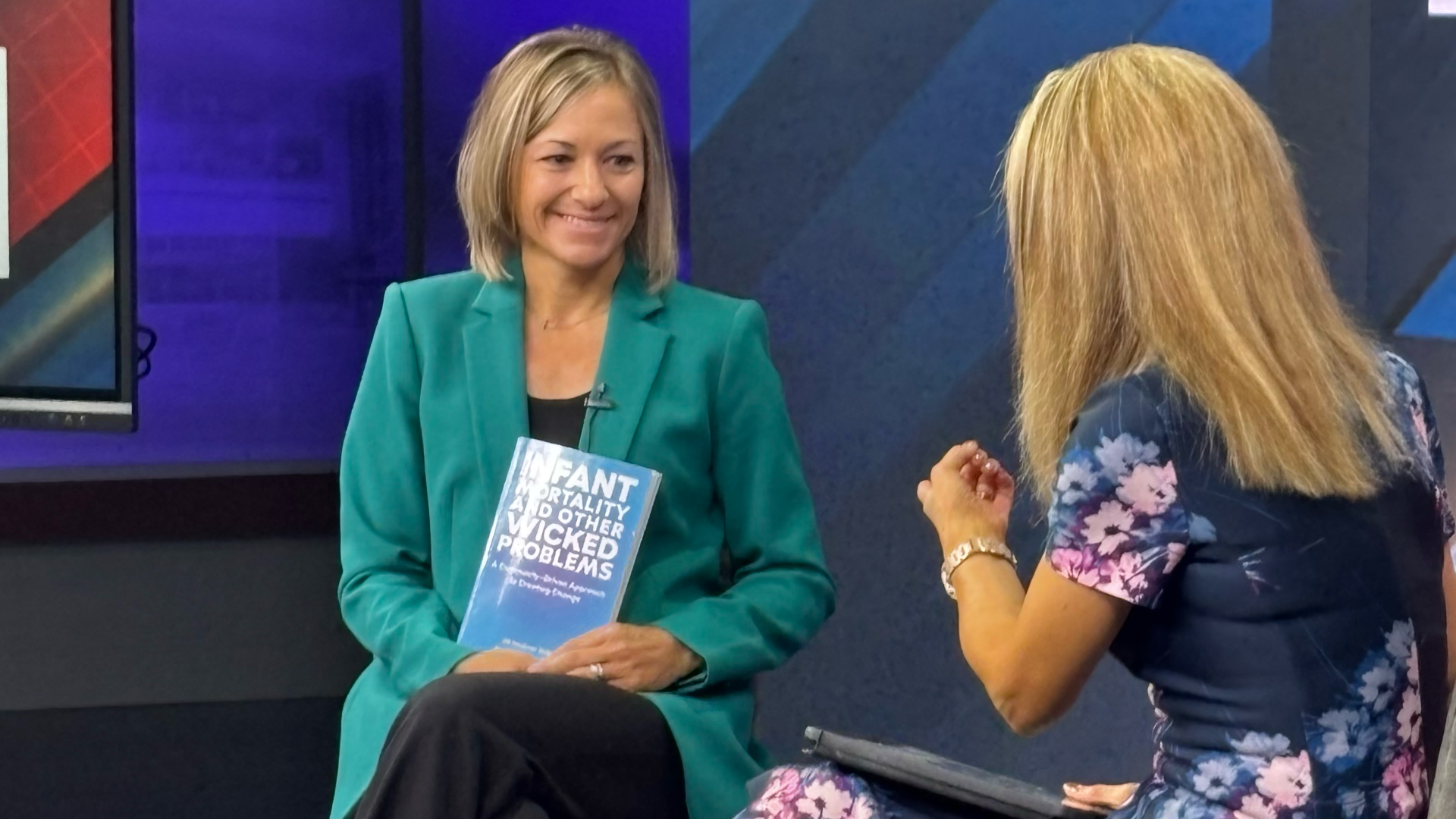 Jill Haubner Miller holding a copy of the book "Infant Mortality and Other Wicked Problems" while talking with Local 12's Liz Bonis